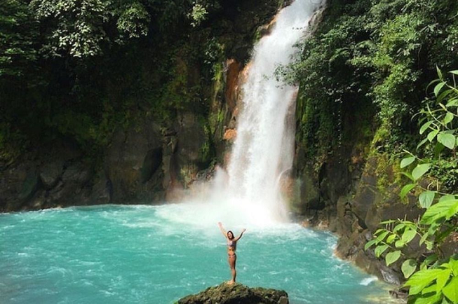 Featured image of top 30 yoga retreats for November 2017 with 8-Day Yoga Adventure, Beach and Volcano Retreat in Costa Rica displaying female yogia with her arms up standing on top of big rock next to shiny blue water and a white roaring waterfall in the background